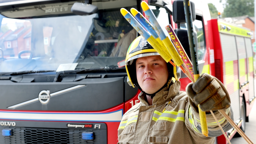 Firefighter Liam Cox holding fireworks while wearing fire kit and standing in front of a fire engine.
